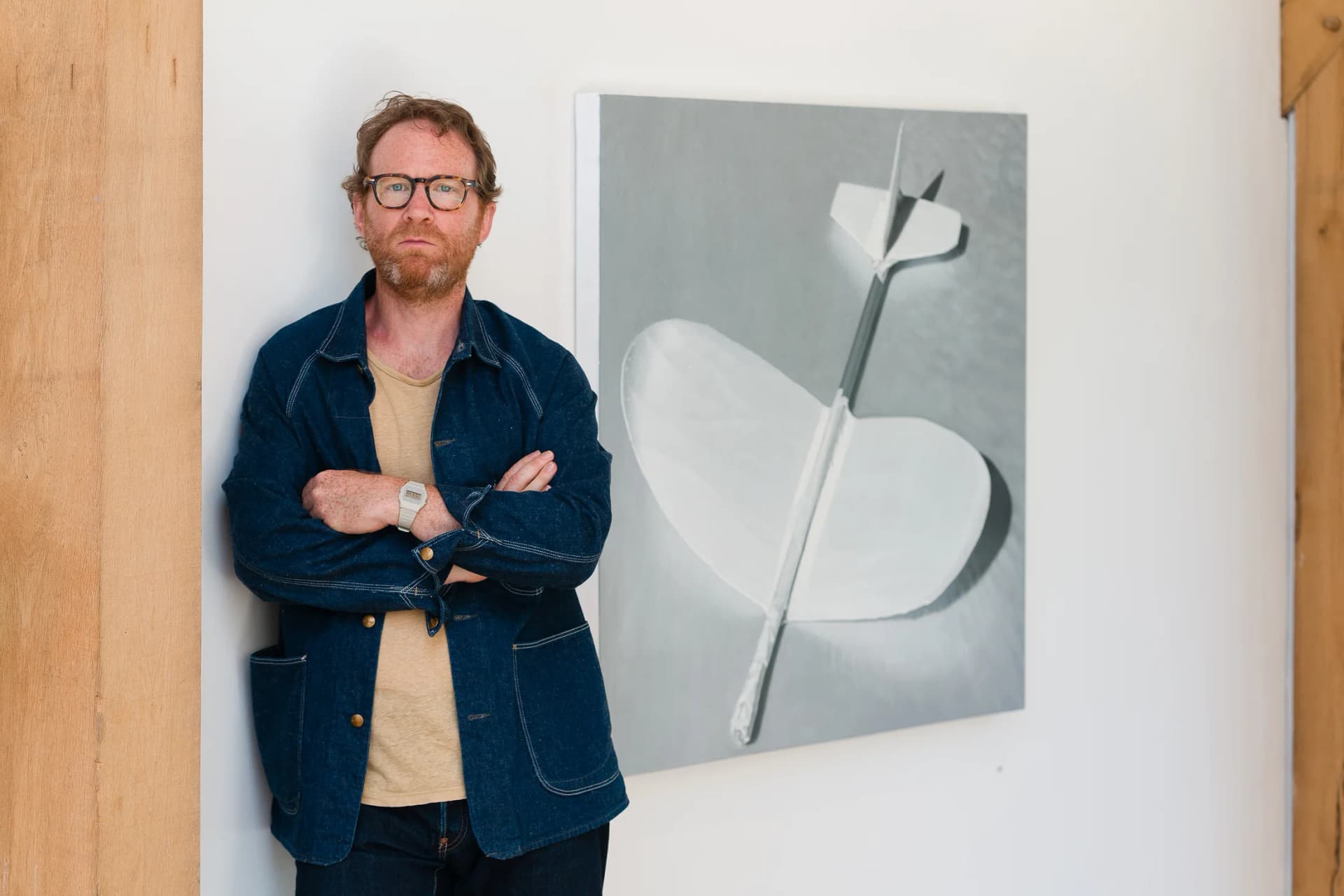 A portrait of Andrew Bryant stood in his studio in front of a monochrome painting of a toy plane