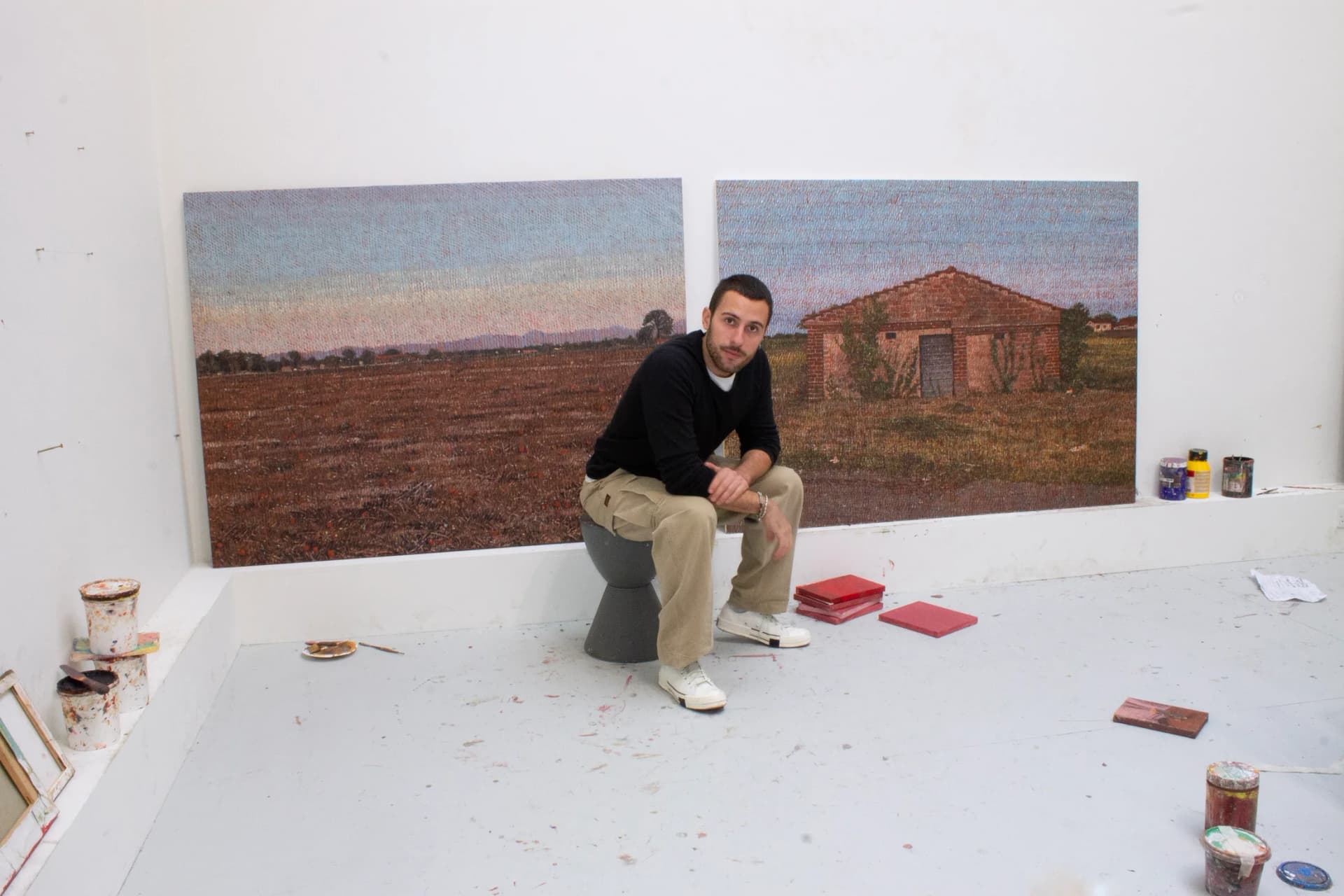 A portrait of Norberto Spina sat in front of two paintings of a landscape and a single storey building