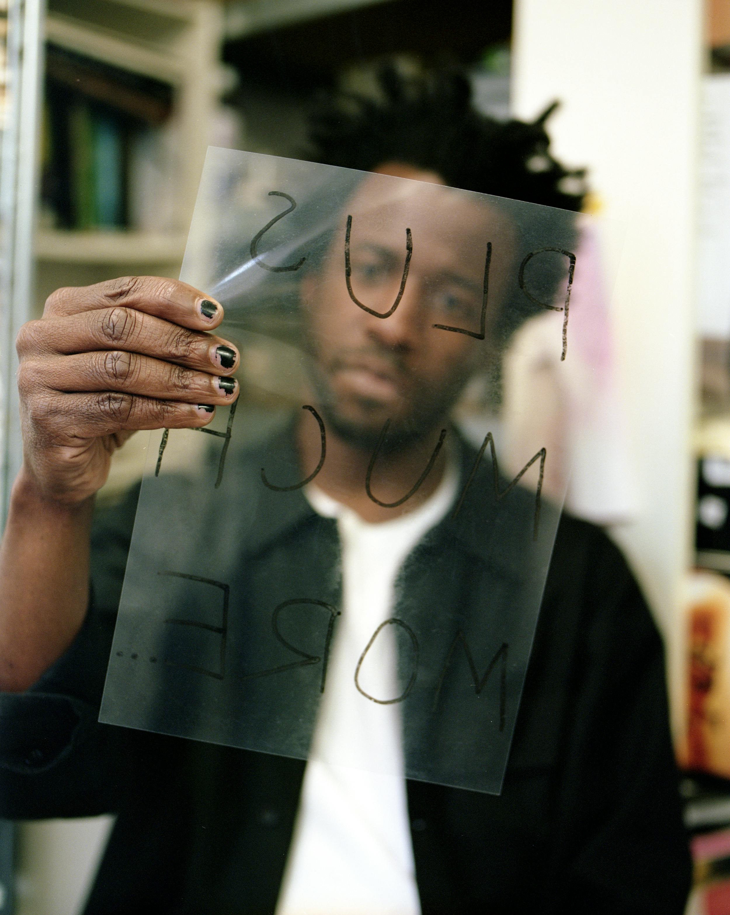 A portrait of Simeon Barclay holding a transparent overhead projector acrylic that says 'plus so much more' backwards in marker pen in front of his face.