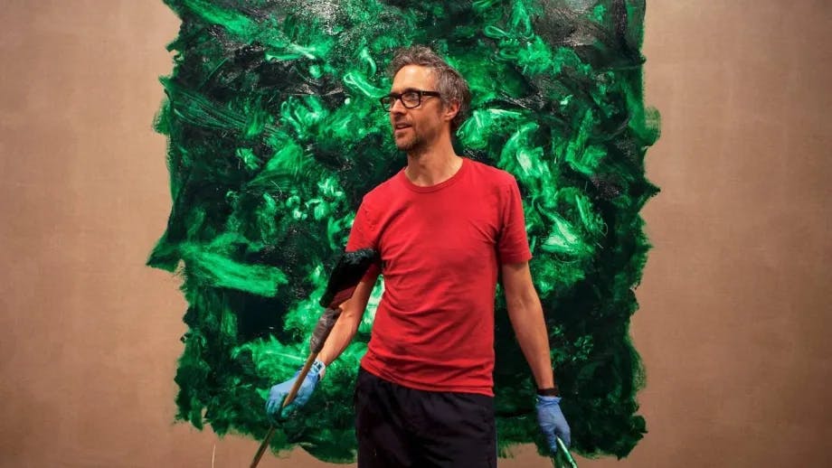 Marcus Coates in front of a swirling green abstract painting wearing a red t-shirt and blue latex gloves